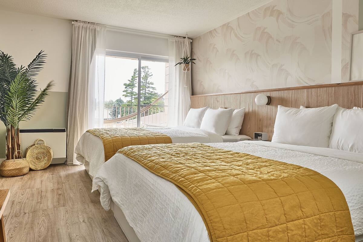 Hotellihuone, jossa on kaksi sänkyä, suuri ikkuna, ruukkukasvi ja näkymä ulos.