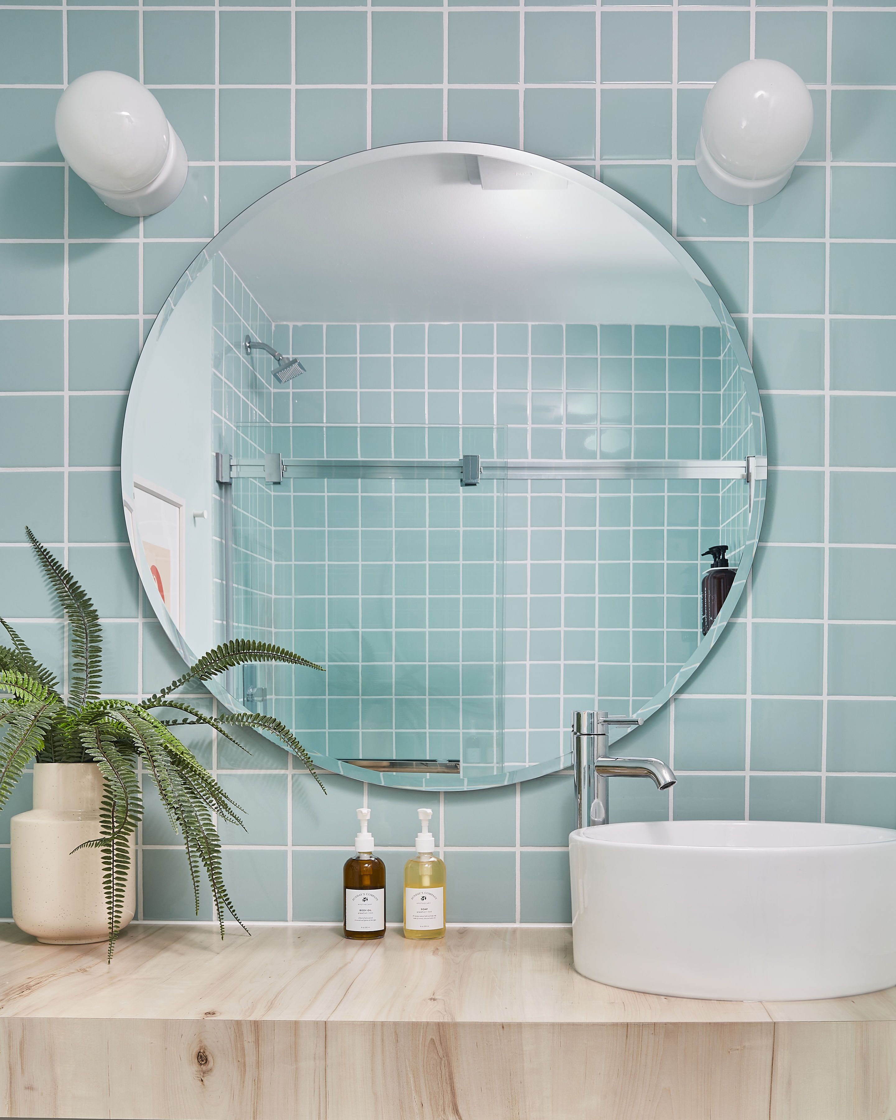 A bathroom with a round mirror, a sink, a shower, and a potted plant.
