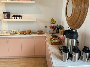 Una cocina con salpicadero de azulejos blancos, un armario rosa, una cesta de frutas y una estación de café con termos etiquetados como 'cream' y 'milk'.