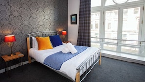 Une chambre d’hôtel avec un lit, deux lampes de chevet, une fenêtre avec des rideaux, un tableau au mur et une vue sur les bâtiments à l’extérieur.
