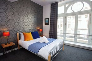 Una habitación de hotel con una cama, dos lámparas de noche, un ventanal con cortinas, un cuadro en la pared y vistas a edificios.