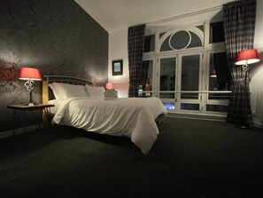 Ein Hotelzimmer mit einem großen Bett, zwei Nachttischen mit Lampen, einem Fenster mit Vorhängen und einer Wand mit einem Musterdesign.