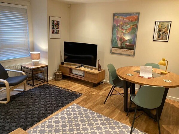 Un salon avec un téléviseur, une table à manger avec des chaises, une lampe et une affiche accrochée au mur.