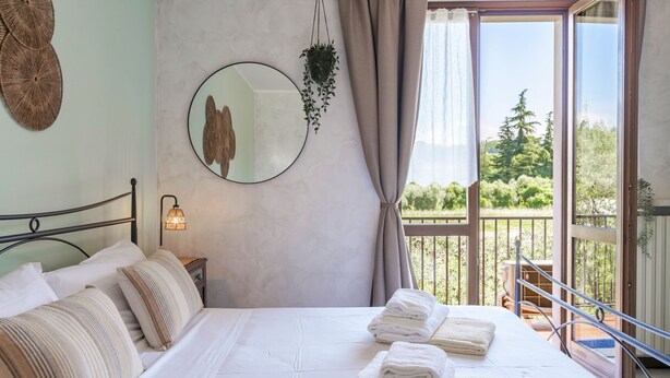 Un dormitorio con cama, almohadas, una planta colgante, un espejo y vistas a zonas verdes por una ventana.