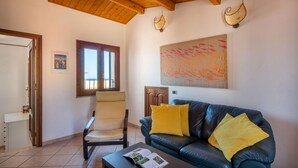 A living room with a leather sofa, a chair, a coffee table with a magazine, a window with a view, and a wall with a textured artwork. - Terrazza Mandralisca by Wonderful Italy (Cefalù)