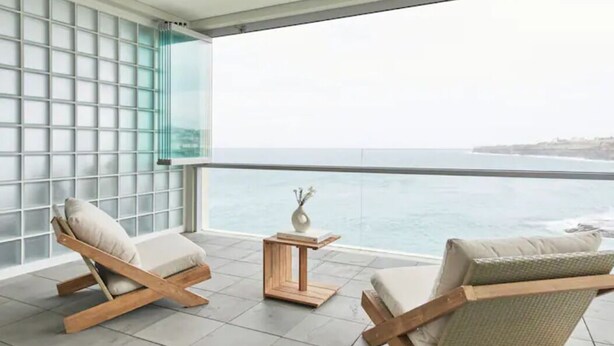 A balcony with two wooden lounge chairs, a small wooden table, and a view of the ocean.