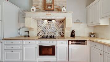 Una cocina con armarios blancos, salpicadero de azulejos, pared de ladrillo, un reloj y una cafetera.
