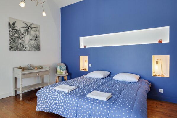 Une chambre avec un mur bleu, un lit recouvert d’une couette à pois blancs, une table de chevet, un bureau et une étagère fixée au mur.