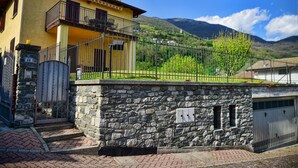 A yellow building with a stone wall and a metal fence, a balcony, and a mountain in the background. - Casa Alex by Wonderful Italy (Gera Lario)