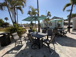 An outdoor patio with tables and chairs, surrounded by palm trees and a building in the background.