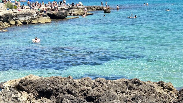 Eine felsige Küste mit klarem, blauem Wasser, Menschen beim Schwimmen und Sonnenbaden, und Sonnenschirme auf den Felsen.