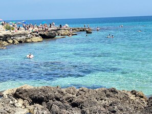 Un litoral rocoso con agua azul cristalina, gente nadando y tomando el sol, y sombrillas sobre las rocas.