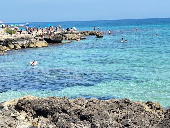 A rocky coastline with clear blue water, people swimming and sunbathing, and umbrellas on the rocks.