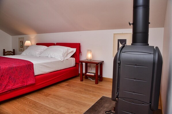 A bedroom with a bed, a red headboard, a bedside table with a lamp, and a black wood stove. - Il Tiglio by Wonderful Italy (Bellagio)