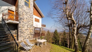 A modern house with a stone facade, wooden balcony, and a paved outdoor seating area with chairs and a table.