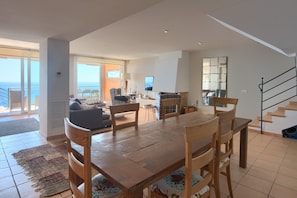 Un vaste espace de vie comprenant une table à manger en bois, des sièges confortables, un téléviseur et une vue sur la mer grâce à de grandes fenêtres.