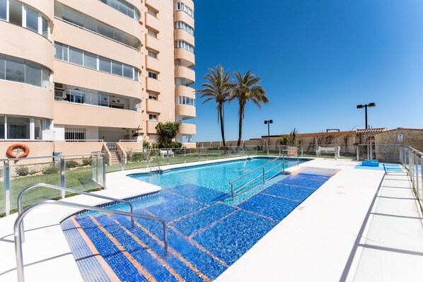 Une piscine dotée d’une terrasse carrelée de carreaux bleus, entourée d’un bâtiment comportant de nombreux balcons et bordée de palmiers.
