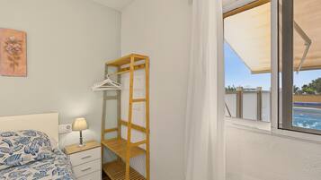 Un dormitorio con cama, mesita de noche con lámpara, estante de madera y ventana con vista a una piscina.
