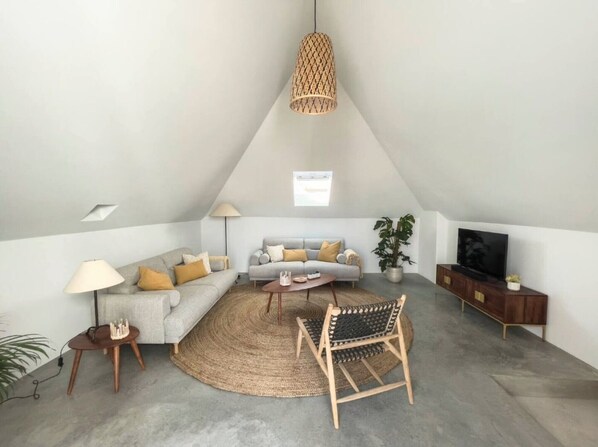 Un salon de style industriel, avec un plafond haut, un luminaire en fibres tressées, un ensemble de canapés, une table basse, un téléviseur et une plante.