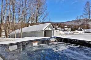 A hot tub with a wooden deck, surrounded by snow-covered ground and trees, with a building in the background. - The Birch Ridge: Family Room #6 - Queen/bunkbed Suite In Killington, Vermont 1 Bedroom Hotel Room (Killington)