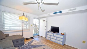 A living room with a sofa, a TV on a blue dresser, a ceiling fan, and a window with blinds. - Old Wharf 15 1 Bedroom Condo by RedAwning (Ocean City)