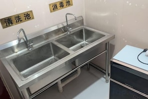 Un lavamanos de acero inoxidable con dos llaves y un dispensador de jabón, instalado en una pared con estructura metálica.