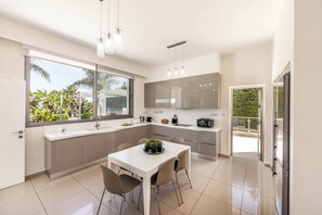 A modern kitchen with a dining area, large windows, and a view of greenery. - Limni Beach Villa - Four Bedroom Villa, Sleeps 8 (Polis)