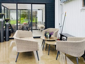 A modern outdoor seating area with wicker chairs and a table, a sliding glass door leading to a garden, and a white wall with a window. - 4 Star Holiday Home in Kallekarr (Kållekärr)
