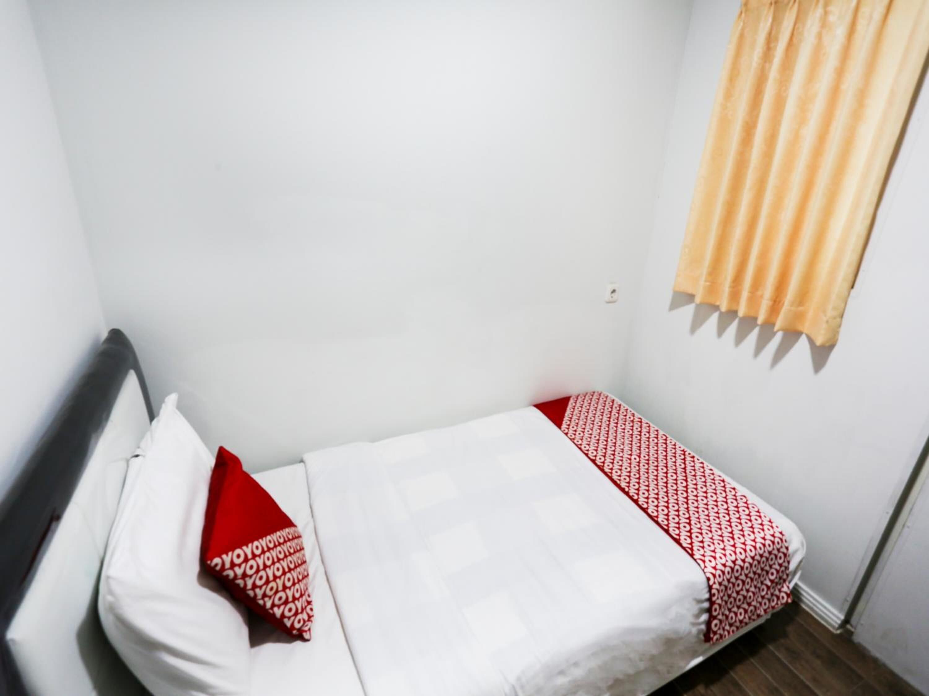 Ein kleines Zimmer mit einem Einzelbett, einer Bettdecke in Weiß und Rot, einem roten Kopfkissen und einem gelben Vorhang.