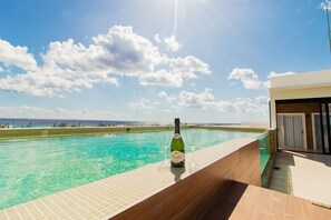 Un espace aménagé au bord de la piscine, avec une bouteille de champagne et deux verres, offrant une vue dégagée sur une piscine aux eaux d’un bleu profond et sur l’océan à l’horizon.