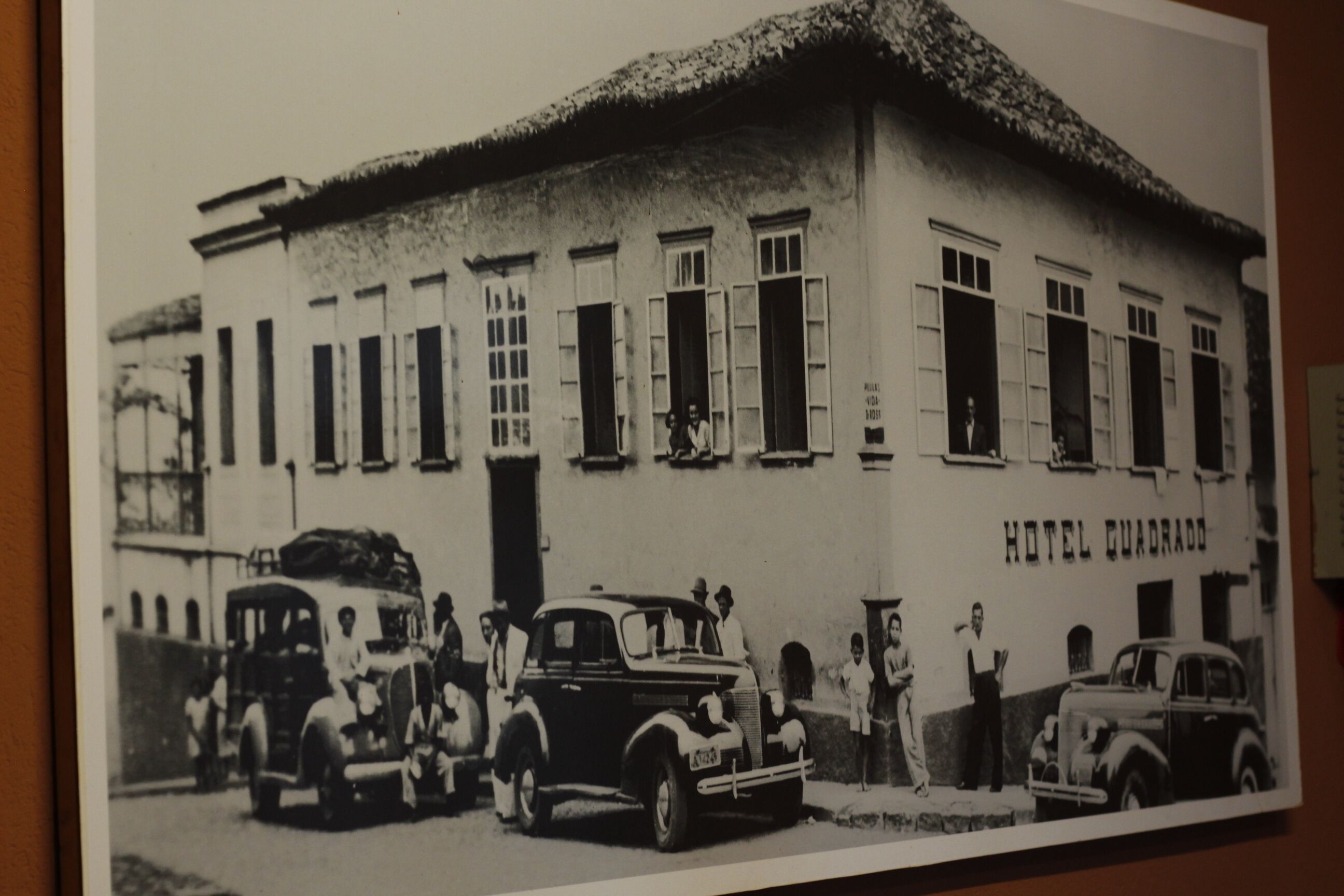 A black and white photo of a building labeled "HOTEL QUADRADO" with vintage cars parked in front and people standing outside.