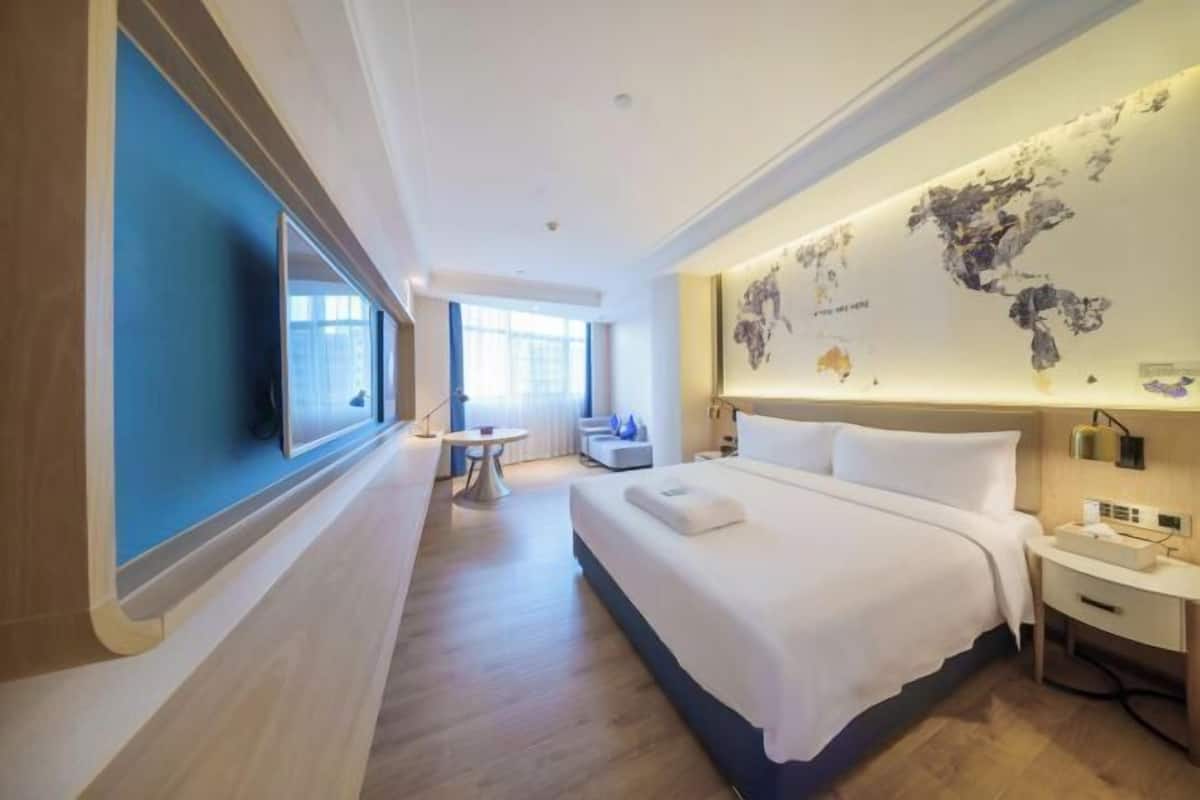 A modern hotel room with a large bed, a desk, a chair, a small table, and a wall-mounted TV.