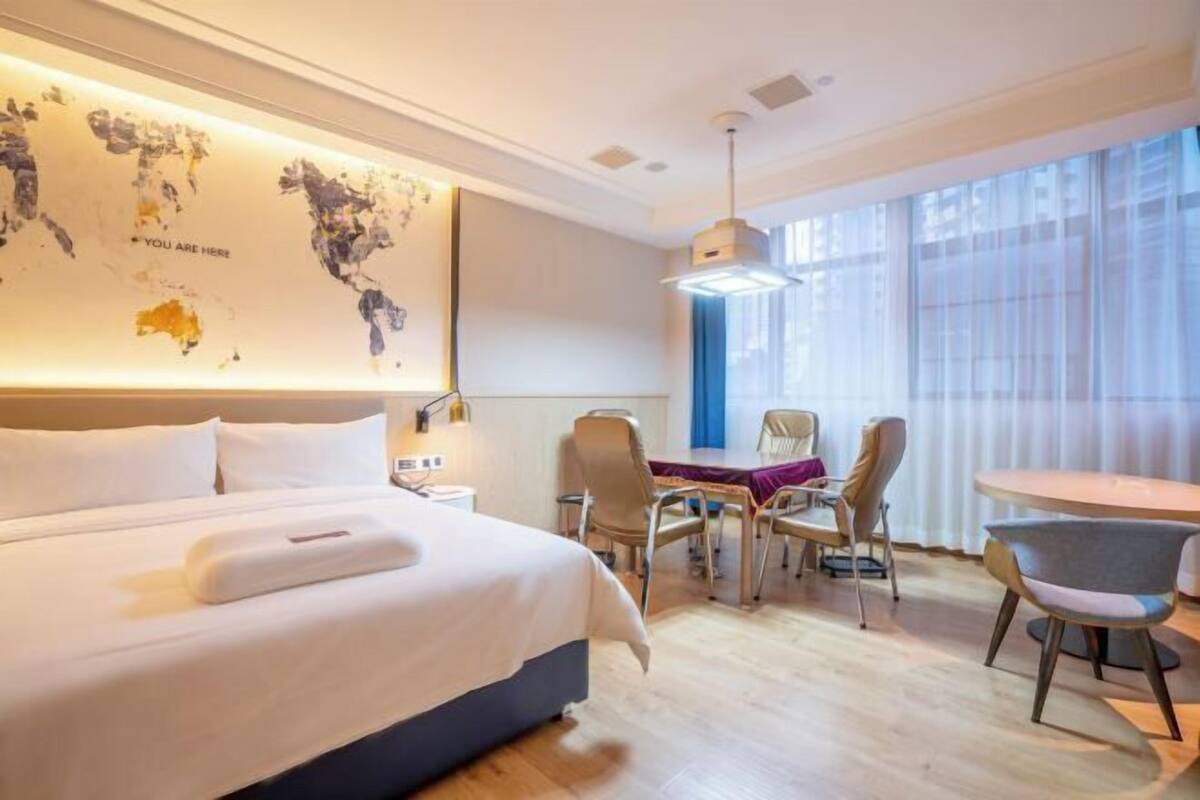 A hotel room with a large bed, a desk with chairs, a map on the wall, and a large window.