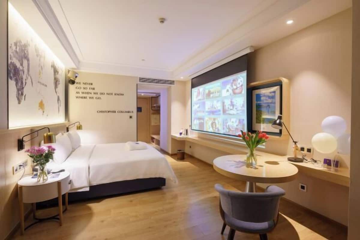 A modern hotel room with a bed, a desk, a chair, a TV screen, and a wall with a quote.