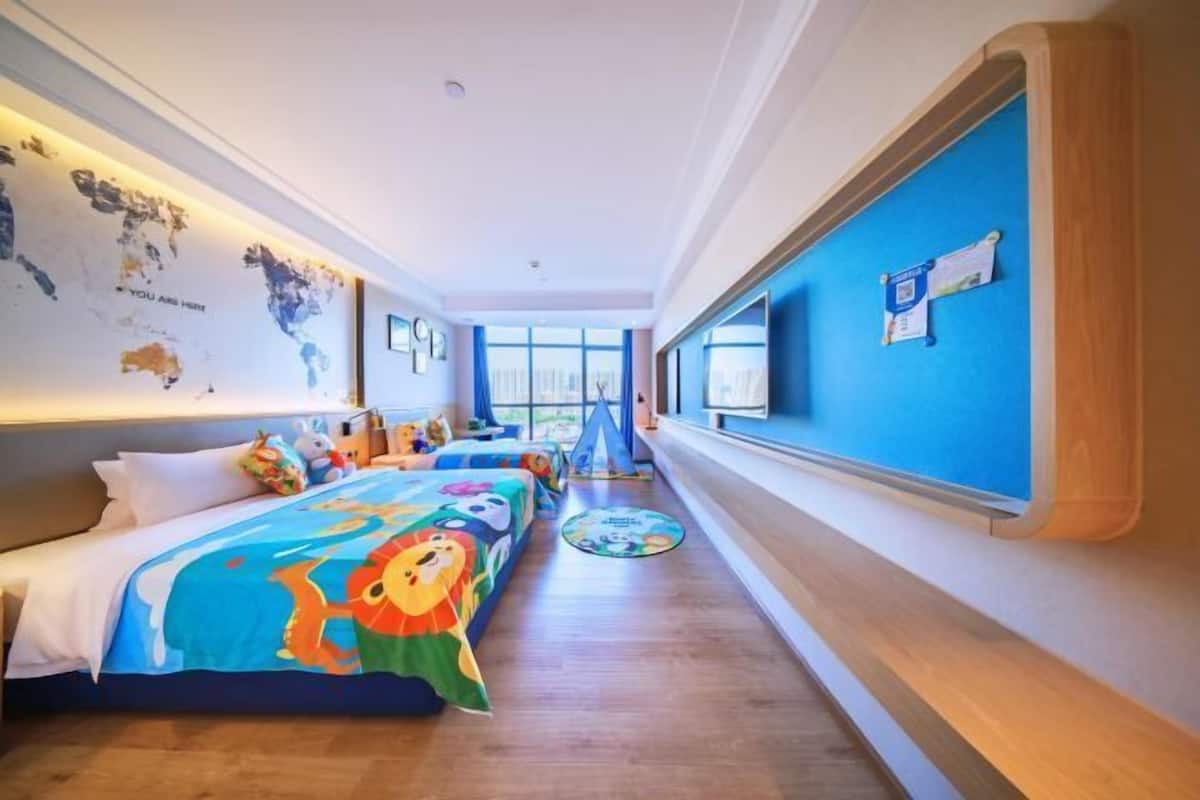 A hotel room with a long bed, a desk, a chair, a window, and a map on the wall.