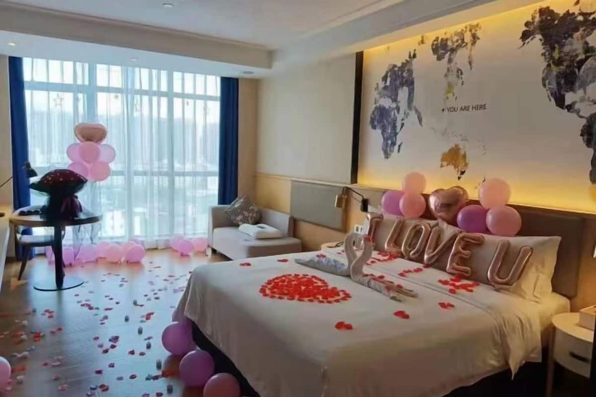 A hotel room with a bed adorned with rose petals and balloons, a world map on the wall, and a view of the city through large windows.