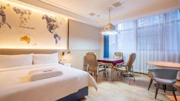 A modern hotel room with a large bed, a desk, and chairs. There is a world map on the wall and a window with curtains.