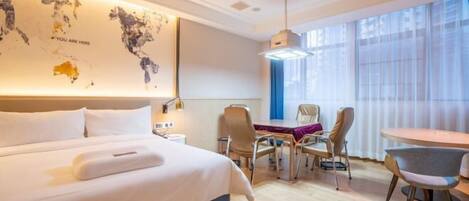A modern hotel room with a large bed, a desk, and chairs. There is a world map on the wall and a window with curtains.