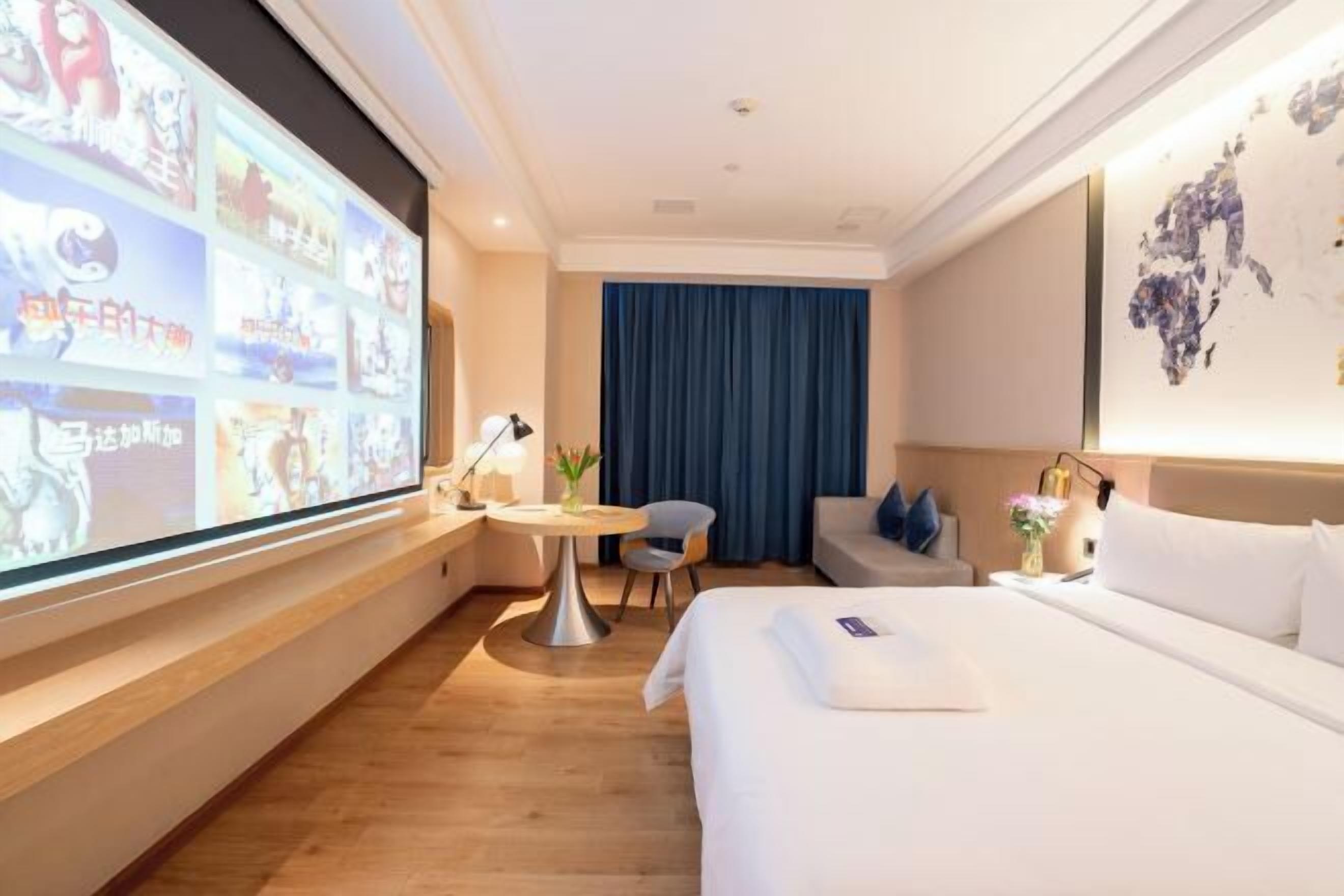 A hotel room with a large screen displaying images, a bed, a desk with a chair, a small table with a lamp, and a blue curtain.