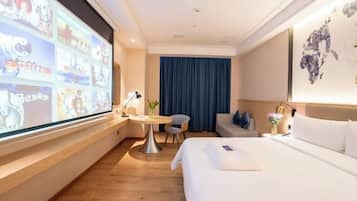 A hotel room with a large screen displaying images, a bed, a desk with a chair, a small table with a lamp, and a blue curtain.