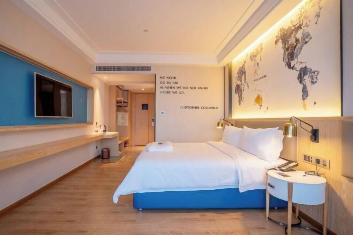A modern hotel room with a large bed, a flat-screen TV mounted on the wall, a world map artwork, and a quote by Christopher Columbus.