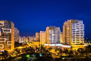 Vista nocturna de una zona residencial con edificios altos, un estacionamiento bien iluminado y árboles.