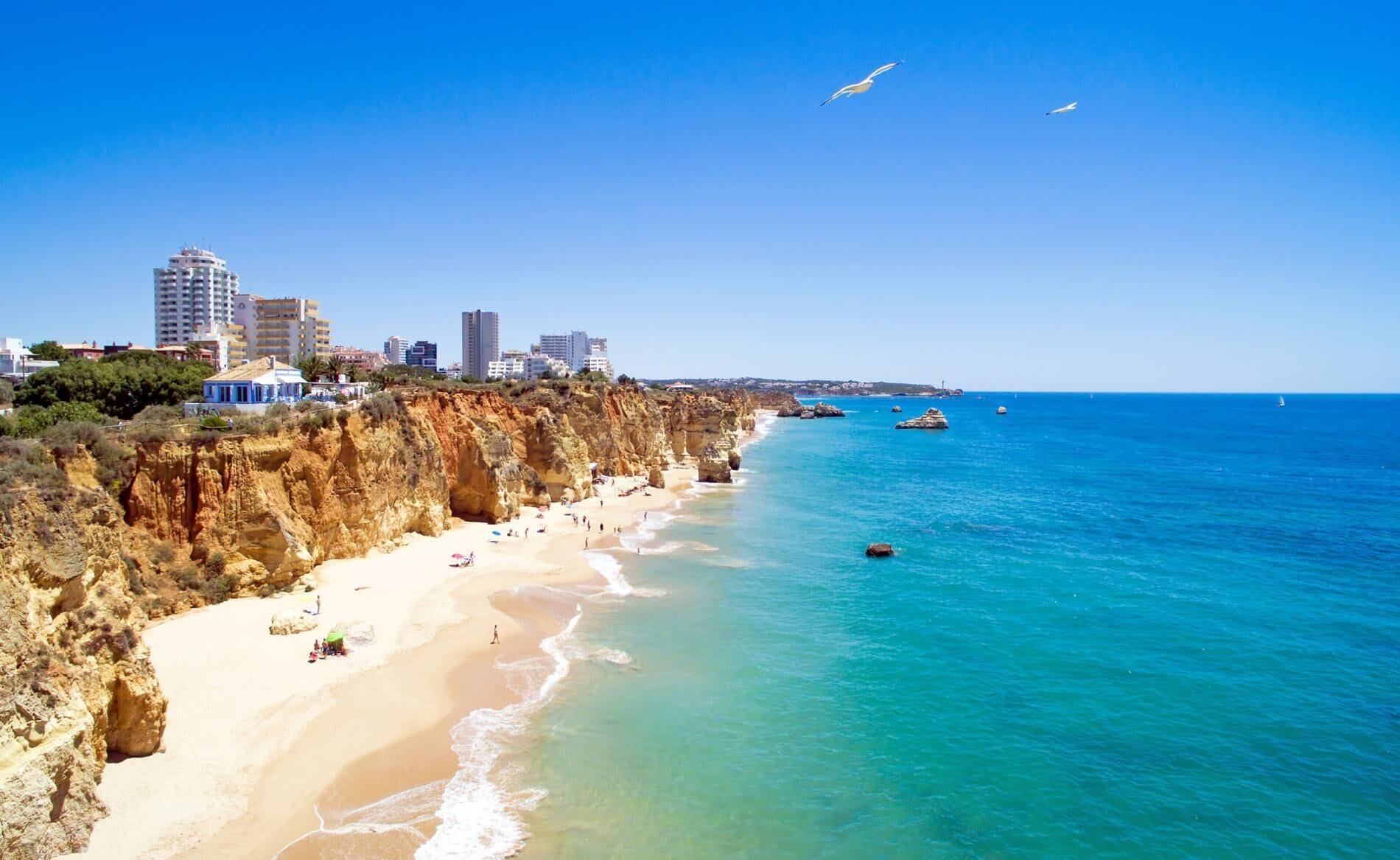 A coastal cityscape with a sandy beach, rocky cliffs, and a clear blue sea.