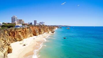 A coastal cityscape with a sandy beach, rocky cliffs, and a clear blue sea.