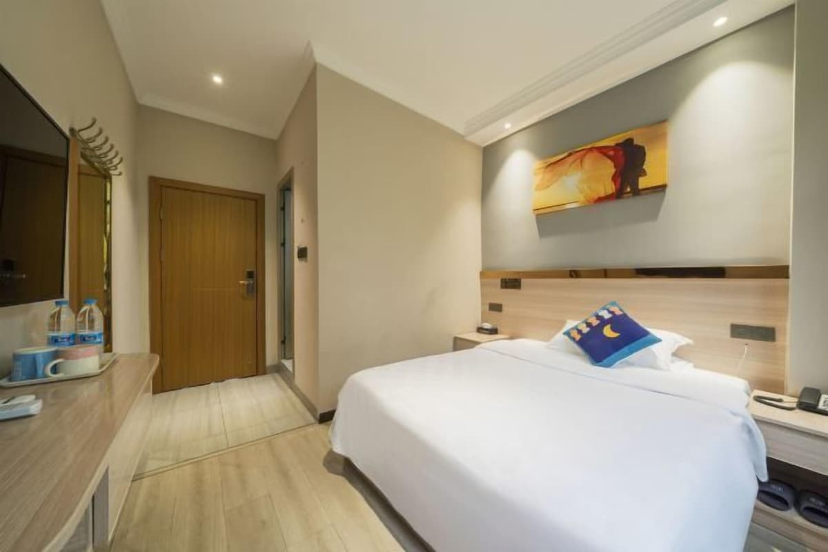 Quarto de hotel com uma cama, uma secretária com cadeira, um espelho, um quadro na parede e uma porta de madeira.