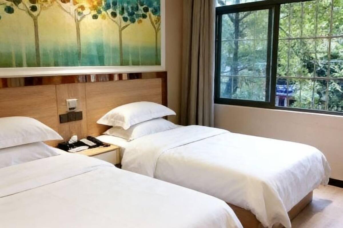 A hotel room with two beds, a nightstand, a painting, and a window with a view of trees.
