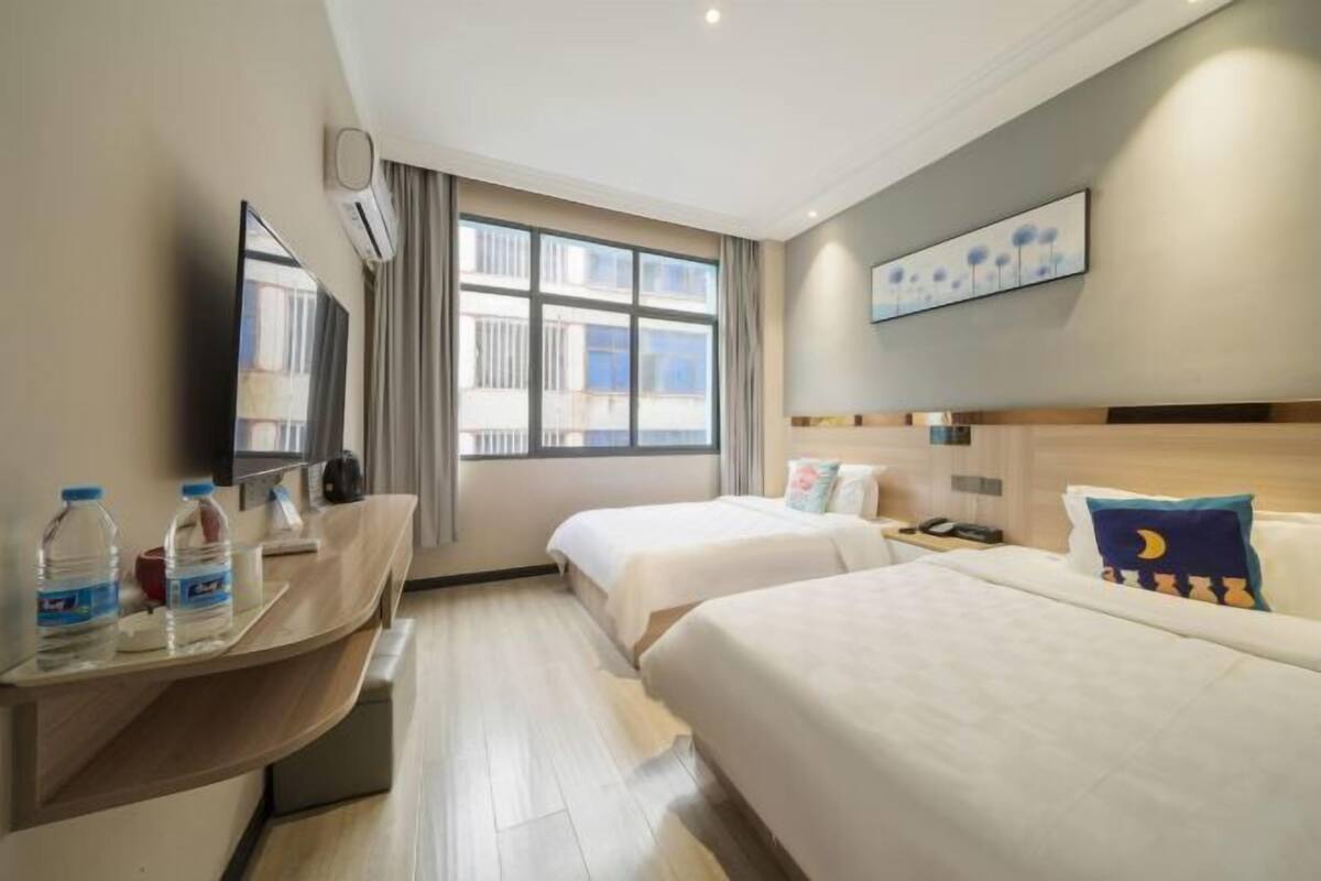 A hotel room with two beds, a desk with water bottles, a mirror, and a window with curtains.