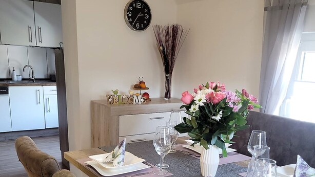 A dining area with a wooden table set for four, surrounded by cushioned chairs. A vase with flowers is on the table, and a clock is on the wall.