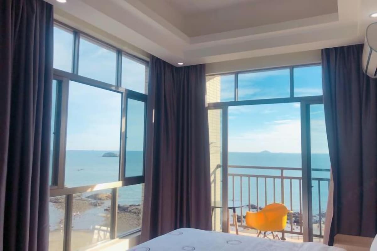 Une chambre d’hôtel avec de grandes fenêtres, un lit, une chaise et un balcon donnant sur la mer.