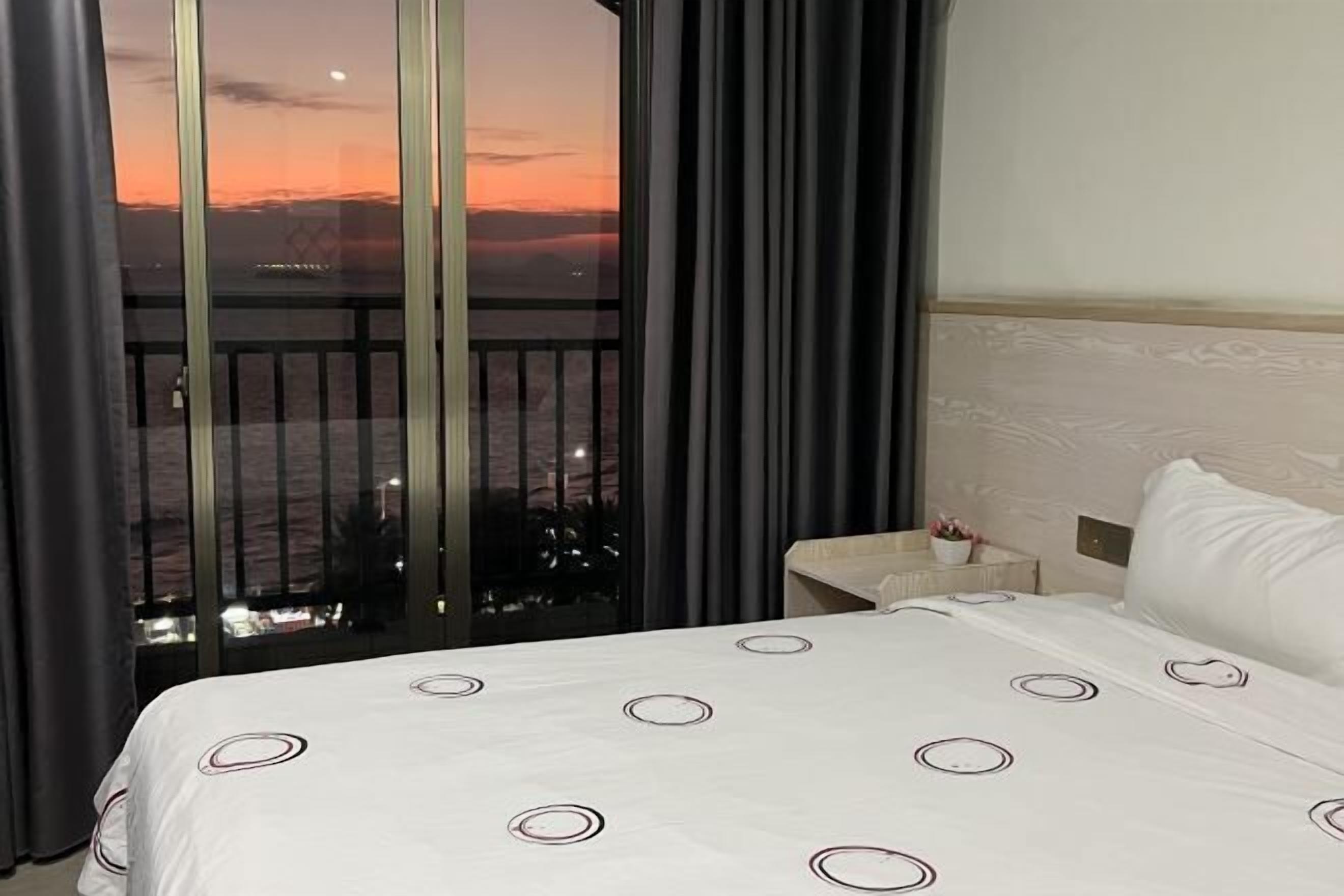 A hotel room with a large bed, a nightstand, and a view of a sunset over a cityscape.